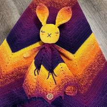 Load image into Gallery viewer, A handmade crochet baby lovey and blanket featuring a yellow bunny with closed eyes, set against a yellow, orange, and purple gradient background, displayed on a grey wood floor.
